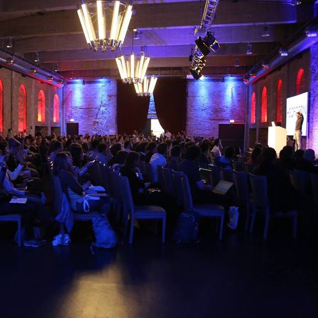 Voll besetzte Industriehalle mit rot beleuchteten Backsteinbögen beim Dataconomy-Vortrag in Berlin